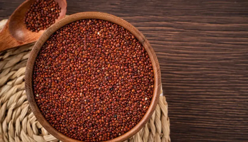 Una ciotola di legno piena di quinoa rossa, con un cucchiaio di legno accanto, su un sottobicchiere intrecciato.