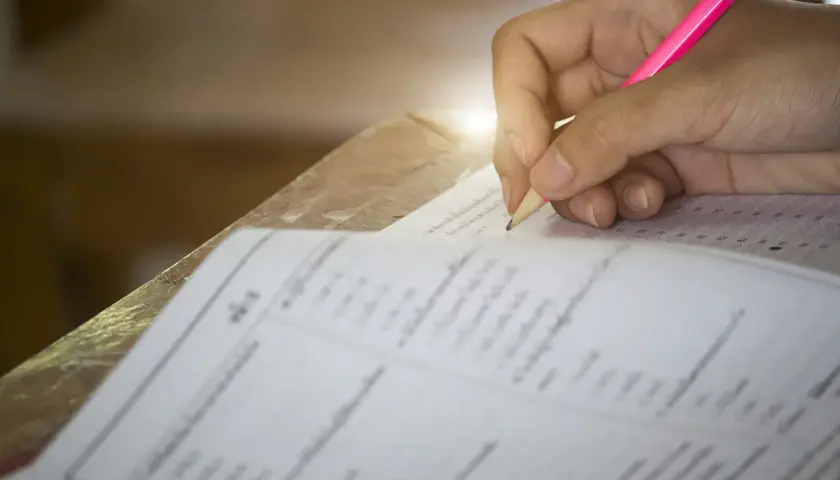 Dłoń ucznia pisząca różowym ołówkiem na arkuszu testowym z kółkami do zaznaczenia odpowiedzi.