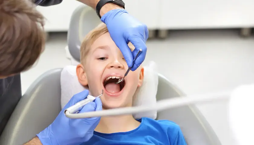 Chłopiec z otwartymi ustami podczas fluoryzacji w szkole. Dentysta w niebieskich rękawiczkach bada ząbki.