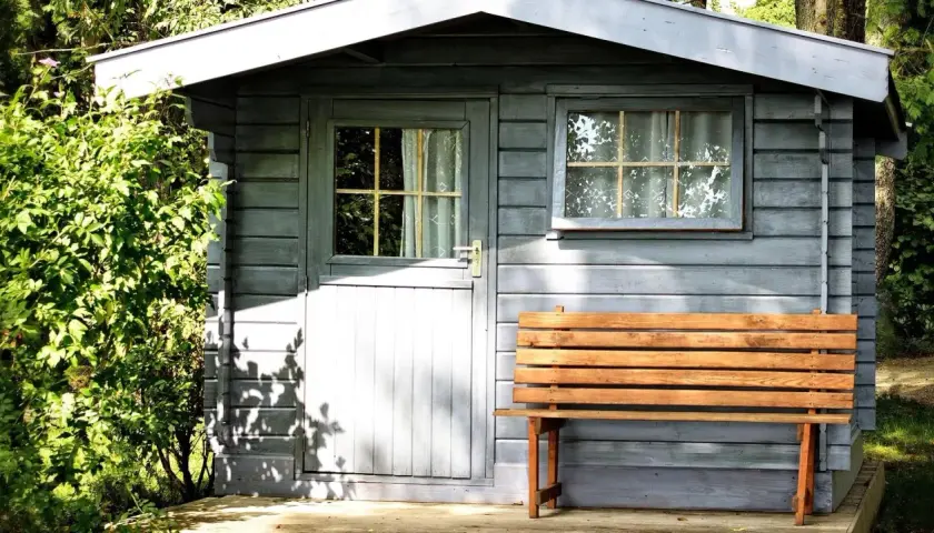 Ein kleines Gartenhaus mit Holzbänken davor. Das Dach ist mit Dachpappe abgedichtet, um es vor Regen zu schützen.