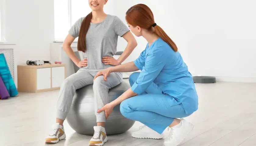 Une kinésithérapeute aide une patiente assise sur un ballon de gym à retrouver de la force dans les jambes grâce à des exercices ciblés.