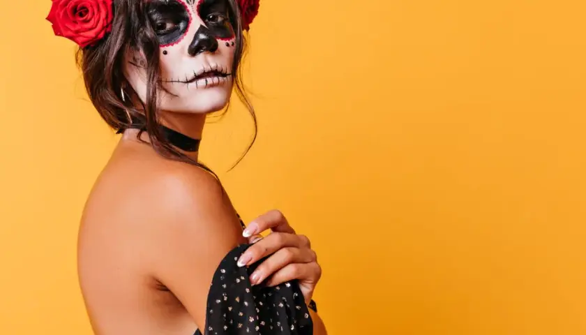 Kobieta z makijażem sugar skull i czerwonymi różami we włosach na pomarańczowym tle.