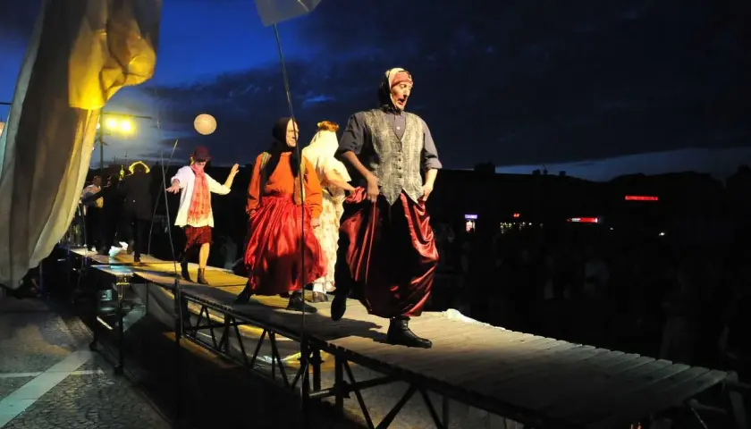 Aktorzy w barwnych strojach na scenie. W tle nocne niebo i widzowie. Spektakl "Teatr ósmego dnia" trwa.