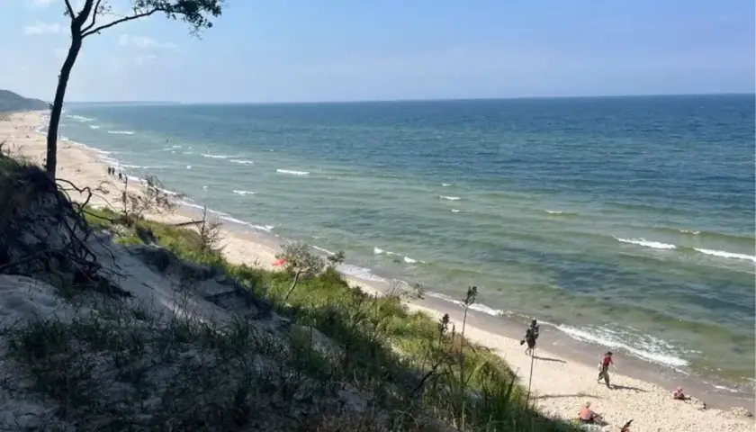 Piękna plaża z wydmami, idealna na orzechowo atrakcje. Woda w morzu jest spokojna, a na piasku widać kilka osób odpoczywających.