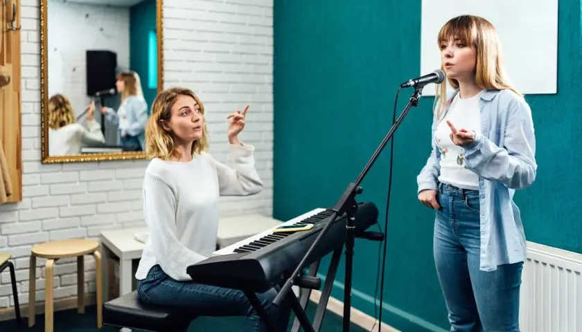 Lekcja śpiewu w domu: nauczycielka przy keyboardzie instruuje uczennicę przed mikrofonem.