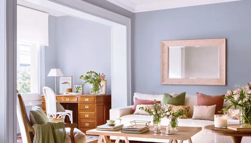 Escritorio de madera con cajonero, silla, sofá con cojines y mesa de centro en una sala de estar luminosa.