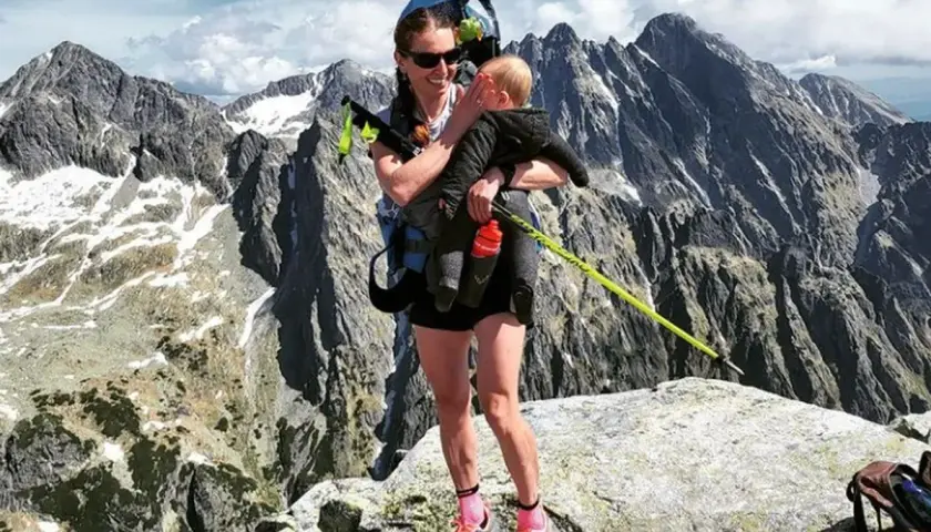 Justyna Kowalczyk z dzieckiem na szczycie górskim, w tle majestatyczne, skaliste pasmo.