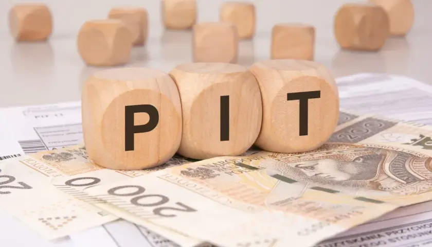 Drewniane klocki z literami "PIT" leżą na banknotach i dokumentach. Może to symbolizować rozliczenie podatkowe lub umowę darowizny.