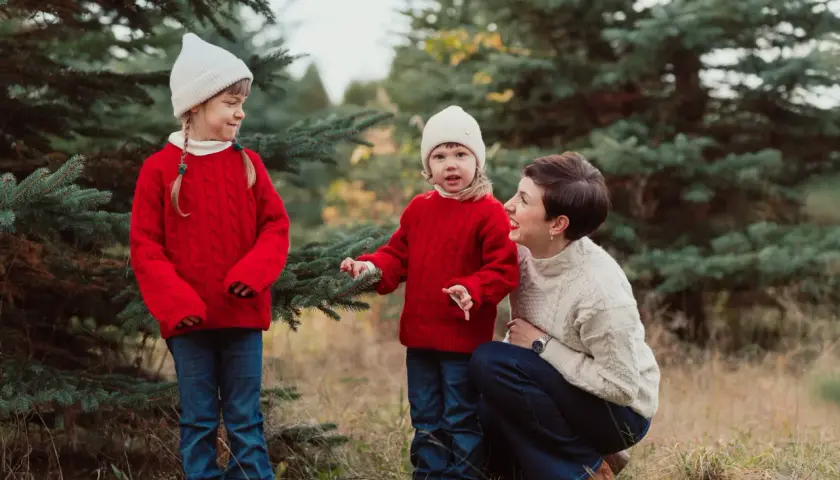 Urocza sesja zdjęciowa dla dziecka w otoczeniu choinek. Mama i dwie córki w czerwonych sweterkach i białych czapkach cieszą się wspólnym czasem.