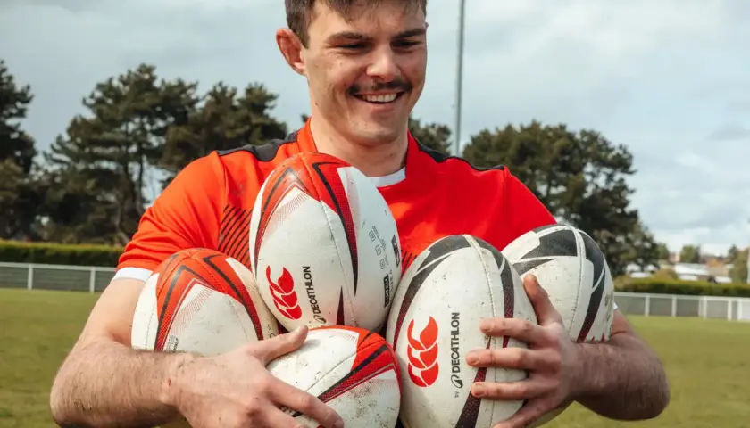 Uśmiechnięty mężczyzna w czerwonej koszulce trzyma kilka piłek do rugby i futbolu amerykańskiego.