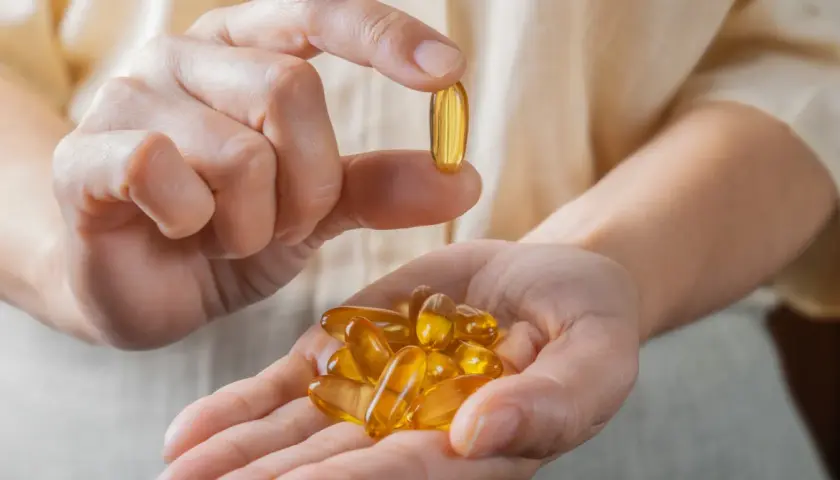 Dłoń trzyma garść kapsułek z olejem rybim, bogatym w kwasy omega 3. Jedna kapsułka jest podniesiona między palcami.