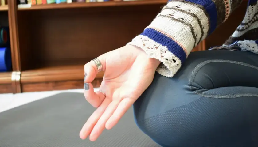 En hand i anjali mudra vilar på ett knä. Bakom syns en bokhylla med yogamattor.