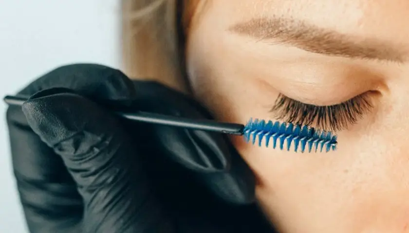 Stylist w czarnej rękawiczce szczotkuje przedłużone rzęsy. Zastanawiasz się, jak długo odrastają rzęsy po przedłużaniu?