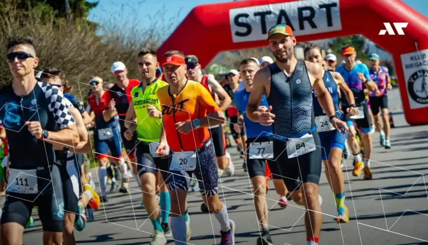 Zawodnicy biegną pod czerwoną bramką startową. Wszyscy przestrzegają zasad triathlonu, skupieni na rywalizacji.