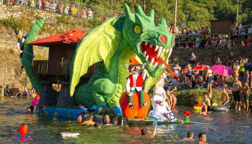 Dragón verde gigante en un desfile acuático, parte de las fiestas de prao Asturias. Gente en el agua y espectadores en la orilla.