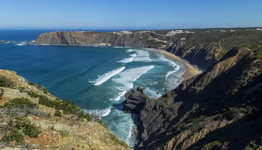 Malownicze wybrzeże Portugalii z klifami, piaszczystą plażą i turkusowym oceanem. Białe fale rozbijają się o brzeg, a w oddali widać białe domki.