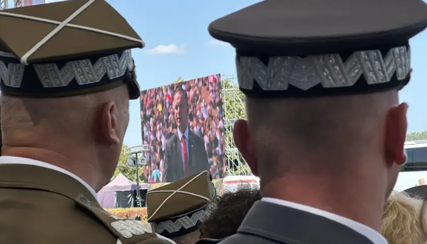 Dwóch mężczyzn w mundurach, jeden z nich ma jedną gwiazdkę na pagonie, ogląda przemówienie na telebimie.