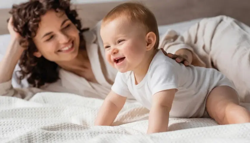 Un feliz bebe 7 meses gatea en la cama mientras su madre sonríe.