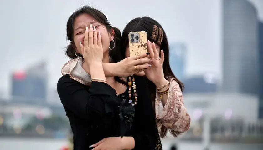 Dwie dziewczyny robią sobie zdjęcie. Jedna zakrywa twarz dłońmi, druga trzyma telefon. W tle widać nowoczesne budynki, jakby z książki o Chinach.