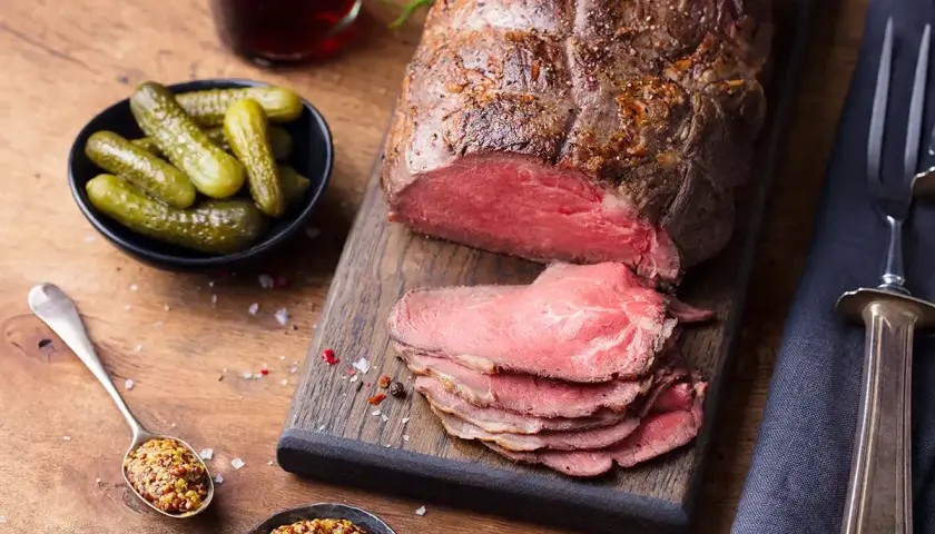 Un rôti de bœuf tranché, servi avec des cornichons, de la moutarde et des couverts sur une planche en bois.