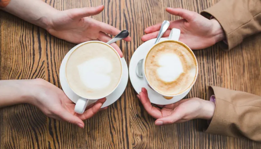 Zwei Cappuccinos mit Herz-Latte-Art, die Hände reichen sie sich über einen Holztisch. Wie viel Milch ist im perfekten Cappuccino?