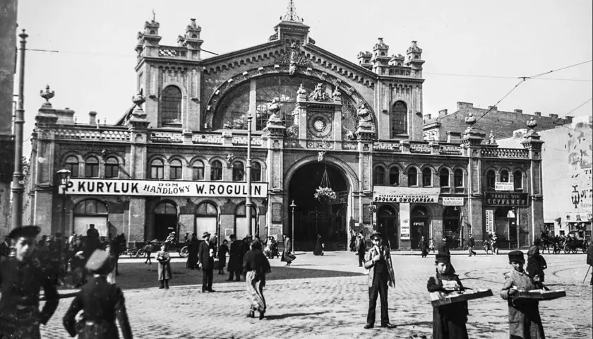 Hala Mirowska tętni życiem. Ludzie spacerują po bruku przed okazałym budynkiem, a dwaj chłopcy niosą tacki z towarami.