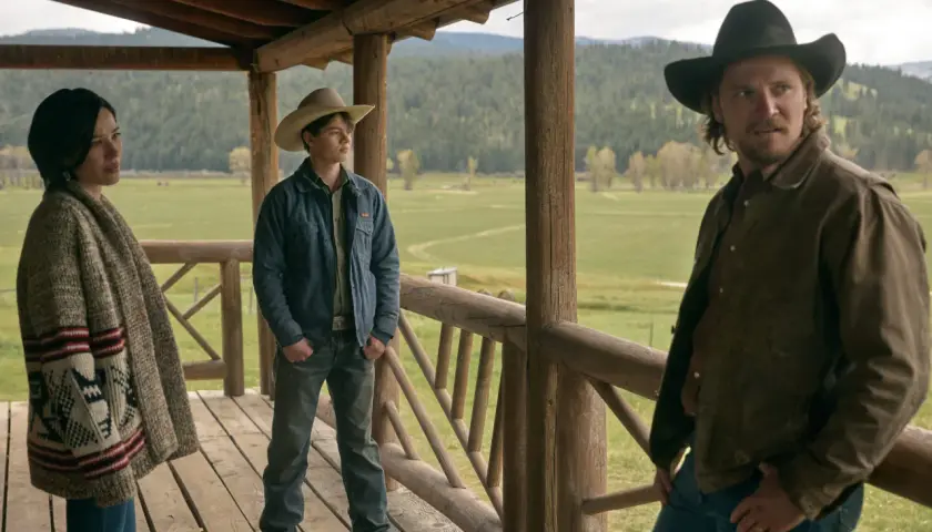 Tre personer på en veranda med berg i bakgrunden. En kvinna med mönstrad kofta, en ung man med cowboyhatt och en man med hatt och brun jacka. Scen från Yellowstone-serien.