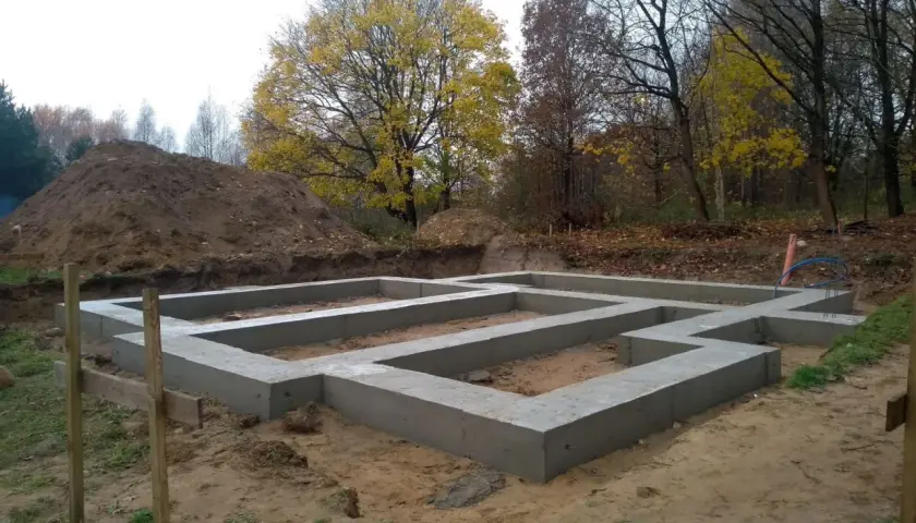 Fundament domu wylewany na ławach fundamentowych, poniżej strefa przemarzania gruntu. W tle jesienne drzewa i usypana ziemia.