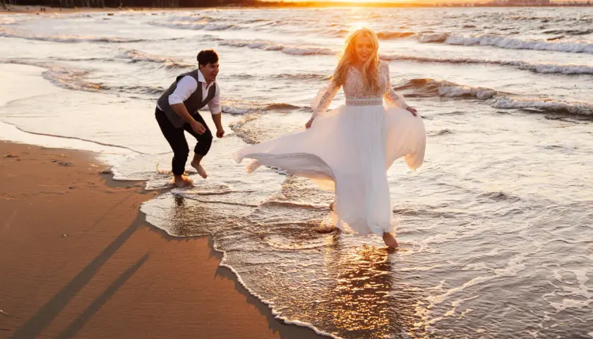 Szczęśliwa para podczas sesji na plaży o zachodzie słońca. Ona w zwiewnej sukni, on w eleganckim stroju, bawią się falami.