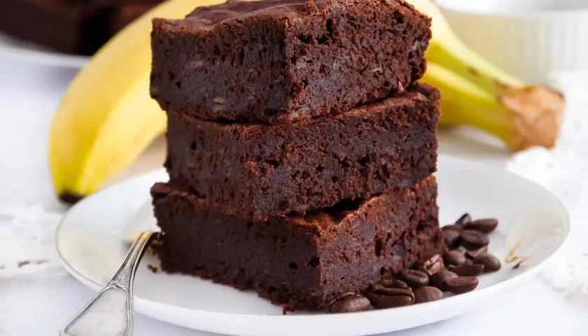 Stos puszystych brownie bananowych na talerzyku, obok dojrzały banan i ziarna kawy.