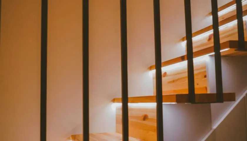 Drewniane stopnie schodów z subtelnym oświetleniem LED w ścianie. Czarne, pionowe balustrady tworzą nowoczesny kontrast.