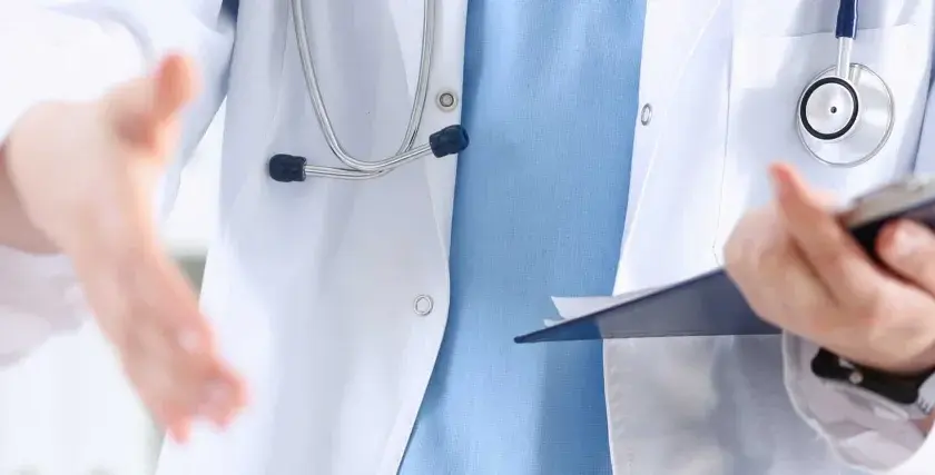 Lekarz w białym kitlu z niebieską koszulą i stetoskopem wyciąga rękę do uścisku, trzymając teczkę. To obraz pracy opiekuna medycznego w hospicjum.