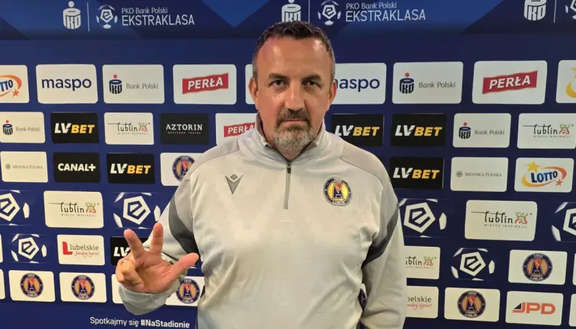 Trener Motoru Lublin z trzema palcami uniesionymi w geście zwycięstwa na tle sponsorów klubu.