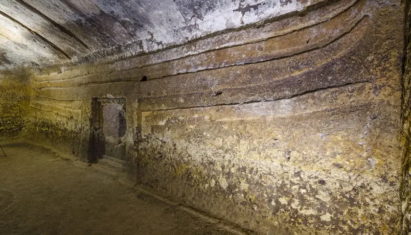 Interno di una domus de janas s'incantu, con pareti scavate e gradini che conducono a un'apertura.