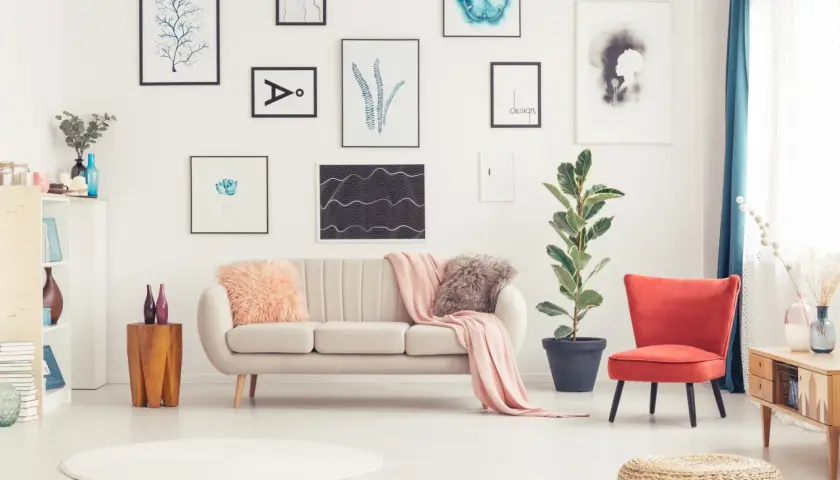 Sofa w kolorze ecru z różowymi poduszkami i pledem, obok czerwony fotel i duża roślina.