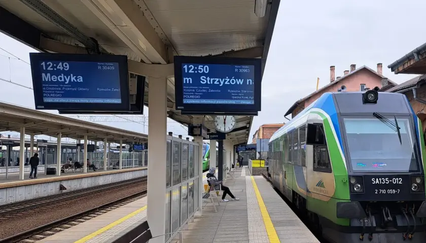 Na peronie kolejowym widać tablice informacyjne z rozkładem jazdy, w tym wskaźnik w4 pokazujący odjazd pociągu do Medyki. Obok stoi zielono-niebieski pociąg.
