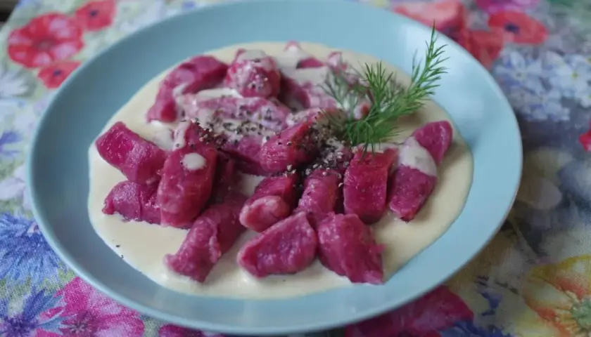 Różowe gnocchi buraczane w kremowym sosie, posypane pieprzem i udekorowane gałązką koperku, na talerzu z kwiecistym obrusem.