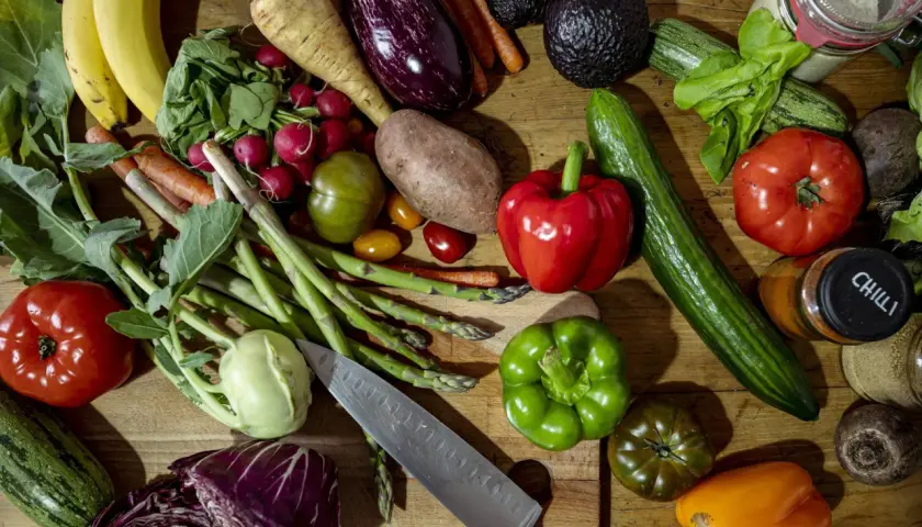 Buntes Arrangement von frischem Gemüse und Obst, darunter Bananen, Paprika, Tomaten, Gurken, Auberginen und Spargel. Ein Messer liegt daneben. Alles für gesundes Essen.