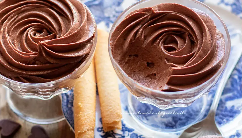 Due coppette di mousse al cioccolato ricetta classica, guarnite con rotoli di wafer e cuoricini di cioccolato.