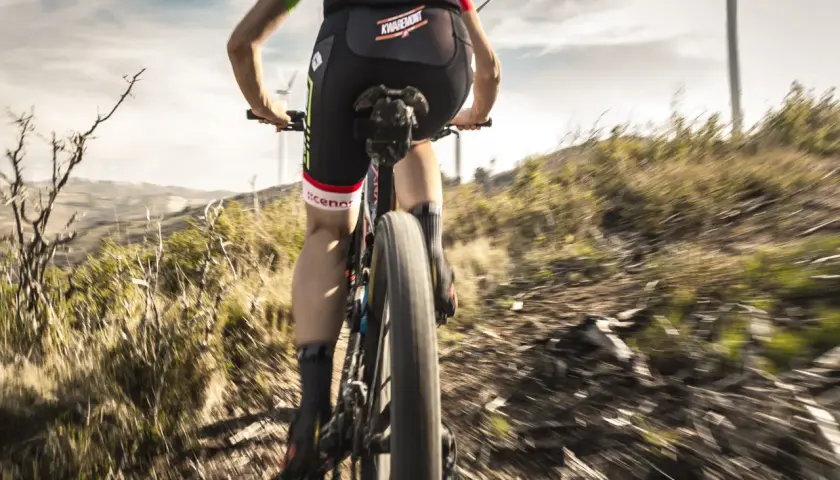 Ciclista desciende por un sendero polvoriento, disfrutando de una de las mejores rutas del mundo en bicicleta. El paisaje árido y las turbinas eólicas al fondo crean una escena épica.
