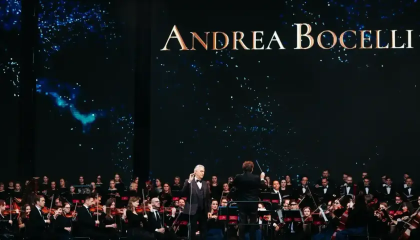 Andrea Bocelli koncert z orkiestrą symfoniczną. Wokalista w garniturze stoi na scenie, dyrygent kieruje muzykami.