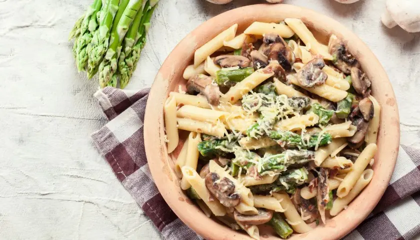 Penne z kremowym sosem, szparagami i pieczarkami, posypane parmezanem. Obok pęczek świeżych szparagów i kilka pieczarek.
