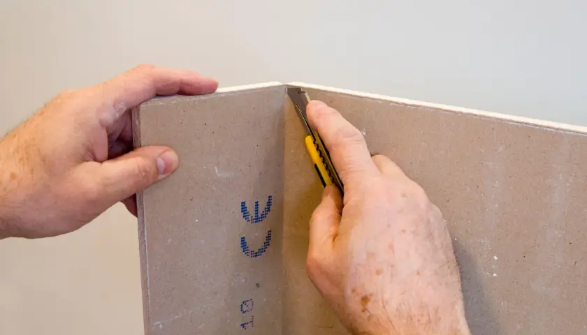 Mani che tengono un cartongesso mentre un taglierino viene usato per tagliare il materiale. Impara come tagliare il cartongesso con precisione.