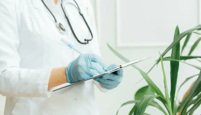 Medico con camice bianco e guanti blu scrive su un blocco appunti. Lo stetoscopio è appeso al collo. Il significato di "extramoenia" è legato a ciò che è fuori dalle mura, come un consulto medico fuori dall'ospedale.