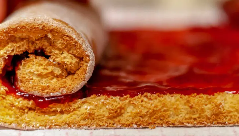 Un rouleau de génoise moelleuse, saupoudré de sucre glace, est en train de rouler sur une couche de confiture de fraises éclatante.
