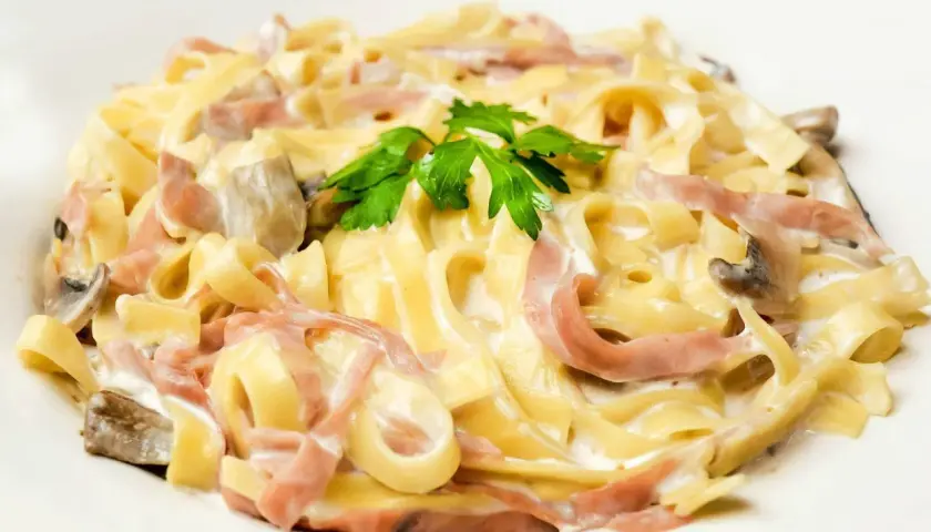 Tagliatelle z szynką i pieczarkami w kremowym sosie, z dodatkiem mozzarelli. Udekorowane natką pietruszki.