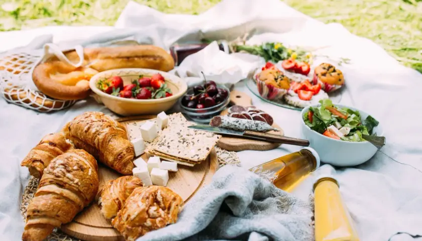 Piknikowy stół z croissantami, owocami, wędliną, sałatką i napojami.