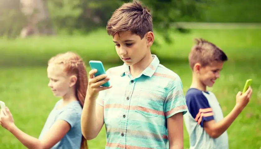 Dzieci w parku korzystają z telefonów. Chłopiec w centrum z niebieskim telefonem, dziewczynka po lewej z białym, a chłopiec po prawej z zielonym. Bezpieczne surfowanie dzięki Google VPN.