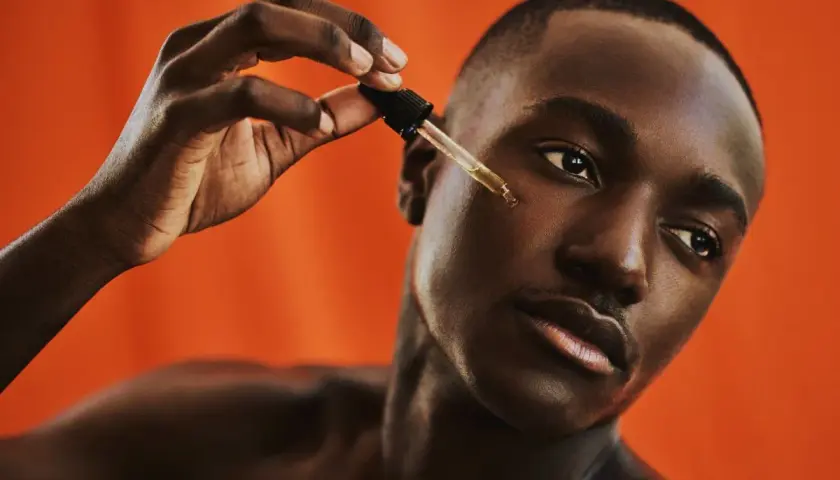 Uomo con pelle scura applica un siero con contagocce, parte della sua beauty routine viso.