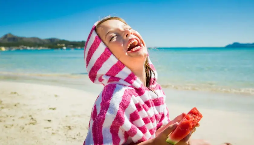 Niña riendo en la playa con una toalla a rayas y sandía, perfecta comida para llevar a la playa con niños.